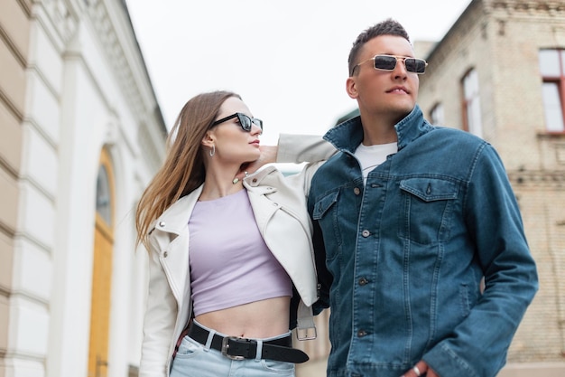 Hermosas y elegantes modelos mujer y hombre con gafas de sol en ropa casual de moda con chaqueta de cuero y chaqueta de jeans caminando en la ciudad