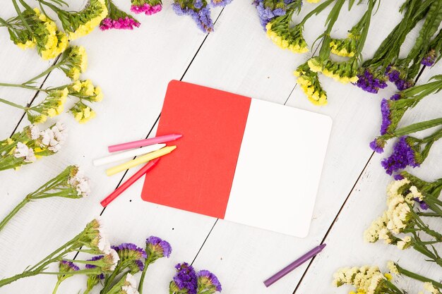 Hermosas flores de colores alrededor de un bloc de notas en blanco abierto con lápices en una mesa de madera blanca