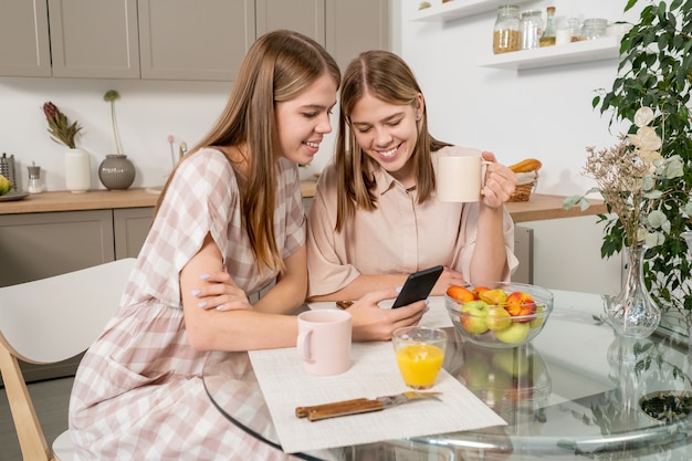 Hermosas hermanas gemelas discutiendo video en el teléfono inteligente mientras están sentados en la mesa de la cocina y tomando bebidas y frutas