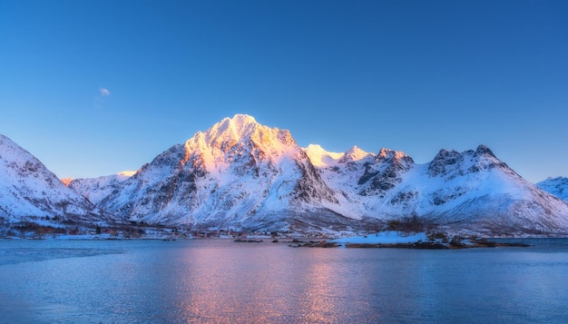 Hermosas montañas cubiertas de nieve y cielo azul reflejado en el agua al atardecer Paisaje invernal con reflejo de rocas nevadas en el mar al atardecer Islas Lofoten Noruega en el crepúsculo Fondo de la naturaleza