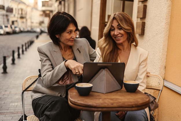 Hermosas mujeres adultas de piel clara se relajan juntas en el café morena muestra tableta a rubia con el dedo Concepto de vacaciones y estilo de vida