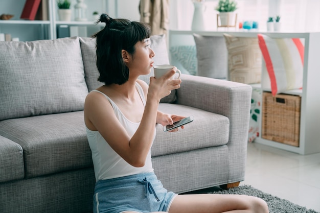 Hermosas mujeres asiáticas bebiendo café con las manos sosteniendo el celular. relájate chica sentada en el suelo en la sala de estar moderna y apoyándose en el sofá mirando hacia el lado. linda mujer disfruta de una taza de té y un teléfono inteligente