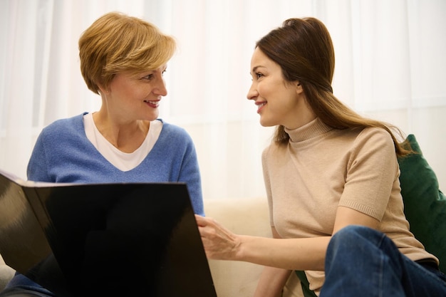 Hermosas mujeres de dos generaciones madre e hija hojeando una revista y hablando mientras se sientan juntas en un sofá en un interior moderno y luminoso