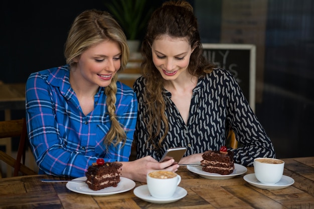 Hermosas mujeres jóvenes con teléfono inteligente con postre y tazas de café en la mesa de café