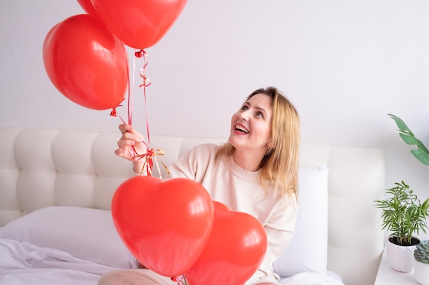 Hermosas mujeres en ropa casual y acogedora en la cama con globos de corazón rojo celebrando el día de san valentín