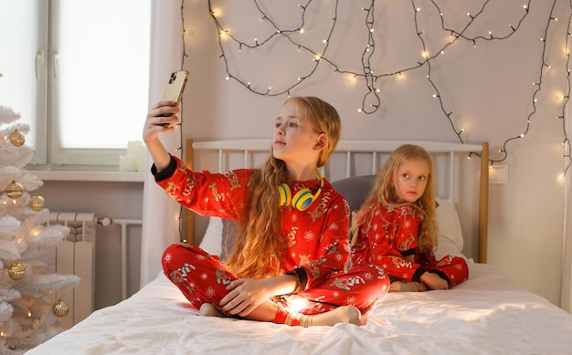 Hermosas niñas están sentadas en la cama con un pijama navideño brillante tomando fotos de sí mismas en el teléfono, una habitación luminosa. Navidad. Año nuevo