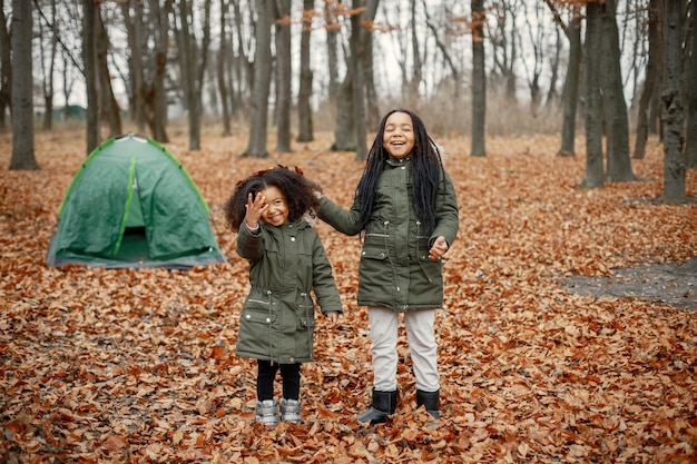 Hermosas niñas negras de pie cerca de la tienda en el bosque Dos hermanitas jugando en el bosque de otoño Niñas negras con abrigos de color caqui