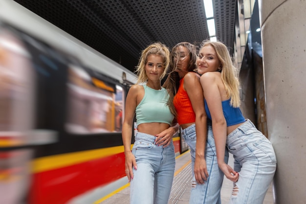 Hermosas novias felices de moda vestidas con ropa informal elegante con puestos de jeans azules y poses en el metro cerca del tren en movimiento