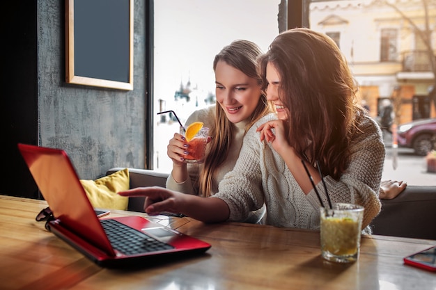 Hermosas y positivas mujeres jóvenes ven películas en la computadora portátil. Ellos sonrien. Modelo sostener vaso de bebida. Otros puntos en pantalla.