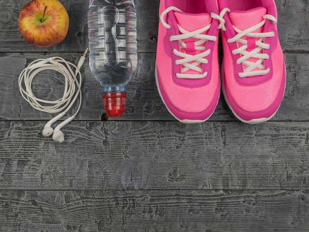 Hermosas zapatillas de color rosa, auriculares, agua y manzanas en un piso de madera. Vista desde arriba.