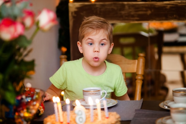 Hermoso y adorable niño de seis años en camisa verde, celebrando su cumpleaños, soplando velas en el pastel horneado casero, interior