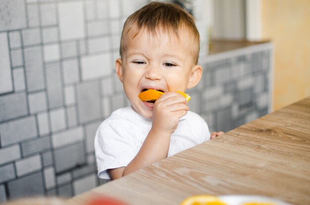 Hermoso Baby Boy en la cocina comiendo ansiosamente una naranja, cortada en gajos