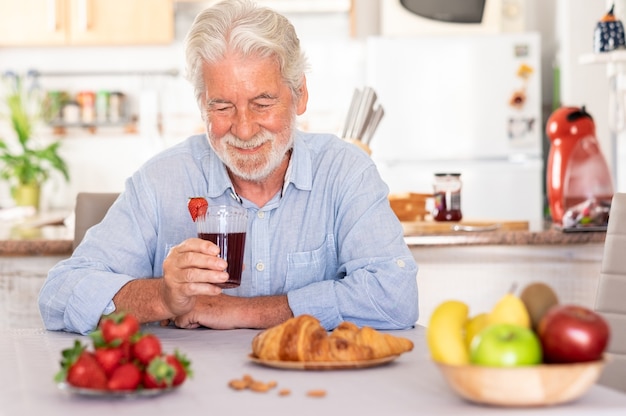 Hermoso cabello blanco senior hombre desayunando en casa, bebiendo un jugo de fresas