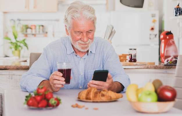 Hermoso cabello blanco senior hombre desayunando en casa mirando las redes sociales en el teléfono inteligente.