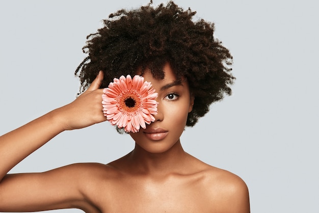 Hermoso como una flor. Atractiva joven africana mirando a la cámara y cubriendo los ojos con una flor mientras está de pie contra el fondo gris