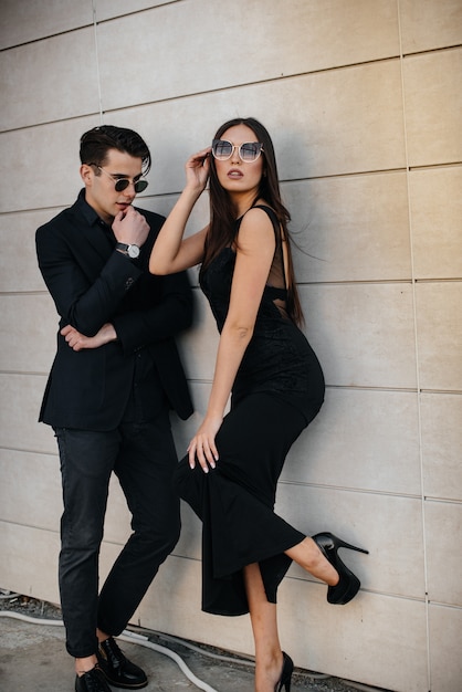 Un hermoso y elegante par de jóvenes vestidos de negro y anteojos se paran contra el fondo de un edificio de oficinas en la puesta de sol. Moda y estilo