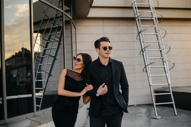 Un hermoso y elegante par de jóvenes vestidos de negro y anteojos se paran contra el fondo de un edificio de oficinas en la puesta de sol. Moda y estilo