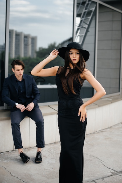 Un hermoso y elegante par de jóvenes vestidos de negro y anteojos se paran en un edificio de oficinas al atardecer. Moda y estilo