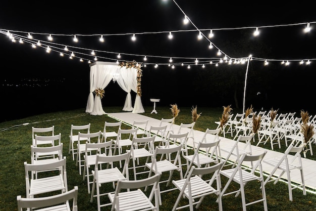 Hermoso escenario nocturno para la ceremonia de la boda. arco de madera de boda. decoración de boda con estilo. sillas para la ceremonia de la boda. bombillas decorativas iluminan el sitio. lugar de boda vacío