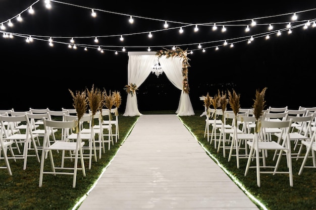 Hermoso escenario nocturno para la ceremonia de la boda. arco de madera de boda. decoración de boda con estilo. sillas para la ceremonia de la boda. bombillas decorativas iluminan el sitio. lugar de boda vacío