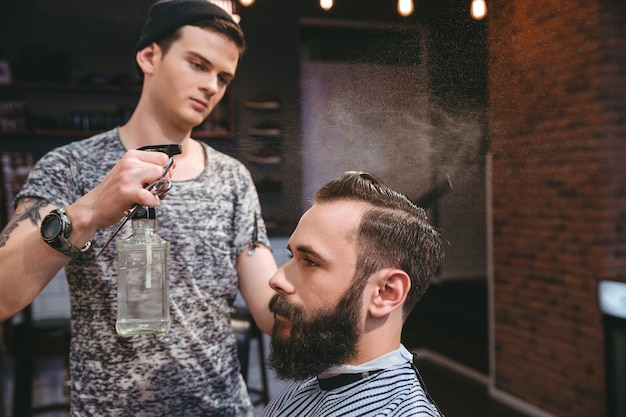 Hermoso estilista concentrado rocía con agua el cabello del atractivo cliente con barba en la peluquería