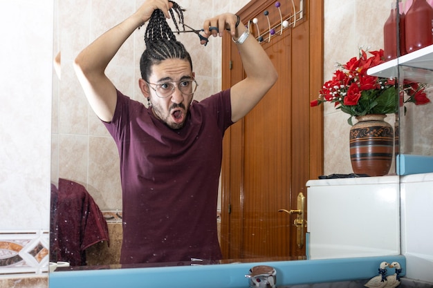 Hermoso hombre asustado mientras se corta el pelo largo en el espejo para donar cabello contra la asociación contra el cáncer