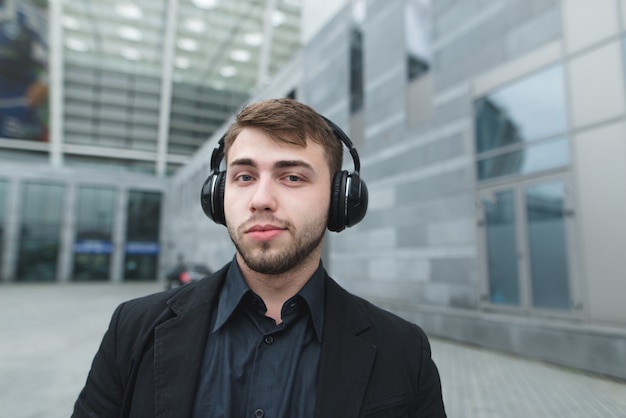 Hermoso hombre con barba y un traje que escucha música en auriculares contra el paisaje urbano.