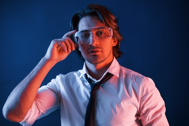 Hermoso hombre con gafas, traje y corbata está en el estudio con luces de neón azul.