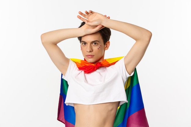 Hermoso hombre gay con brillo en la cara, vistiendo crop top y bandera lgbt arco iris, posando sobre fondo blanco.