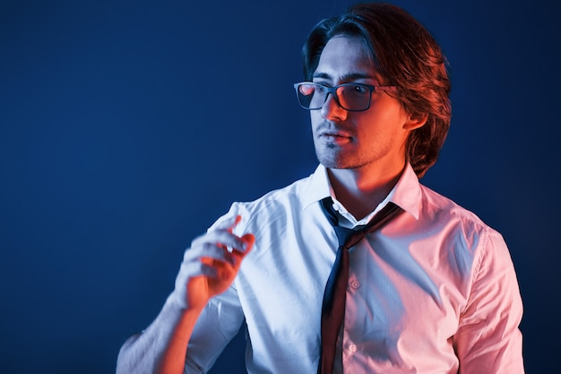 Hermoso hombre con ropa formal y gafas está en el estudio con iluminación de neón azul.