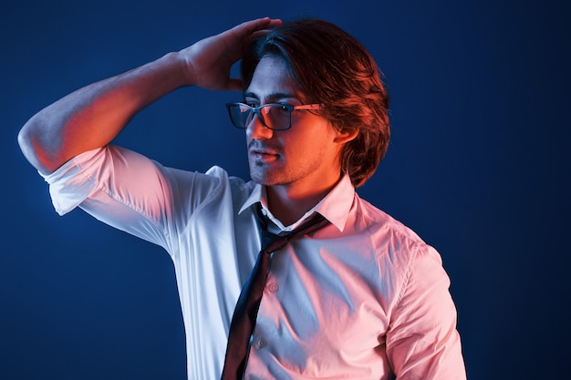 Hermoso hombre con ropa formal y gafas está en el estudio con iluminación de neón azul.