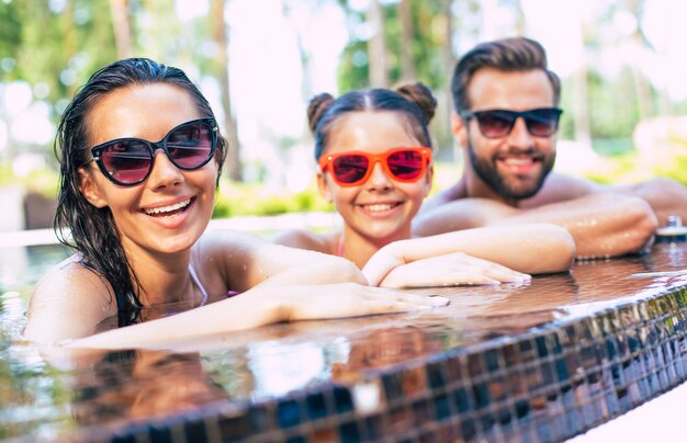 Hermoso hombre sonriente y linda mujer emocionada con su pequeña hija encantadora se divierten en la piscina de verano mientras descansan en el hotel.