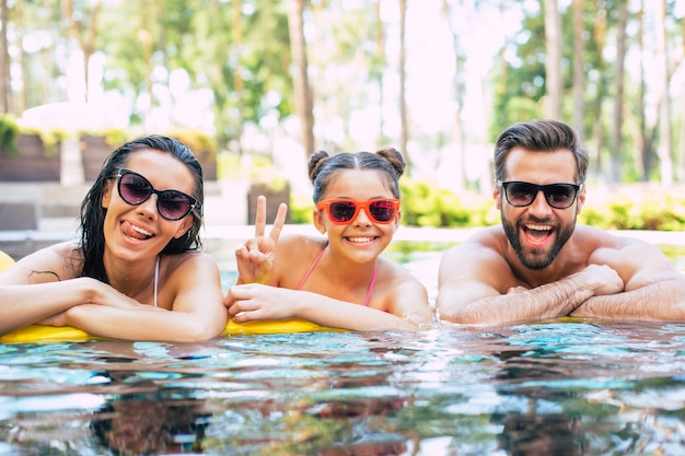 Hermoso hombre sonriente y linda mujer emocionada con su pequeña hija encantadora se divierten en la piscina de verano mientras descansan en el hotel.