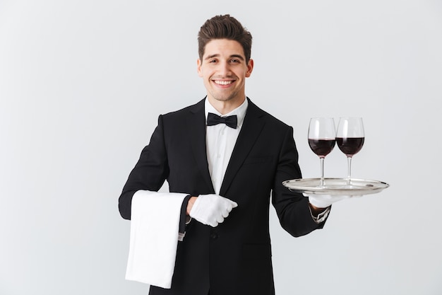 Foto hermoso joven camarero vistiendo esmoquin presentando una bandeja con dos copas de vino tinto aislado sobre pared gris