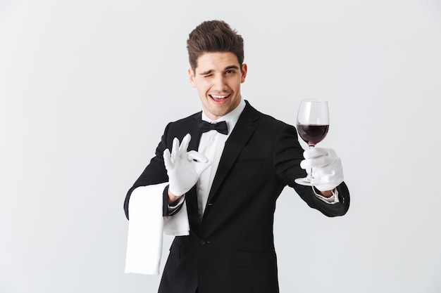 Hermoso joven camarero vistiendo esmoquin sosteniendo una copa de vino tinto aislado sobre pared gris