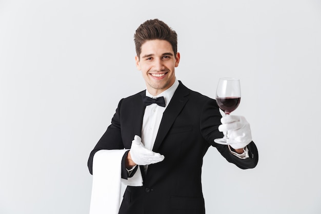 Hermoso joven camarero vistiendo esmoquin sosteniendo una copa de vino tinto aislado sobre pared gris