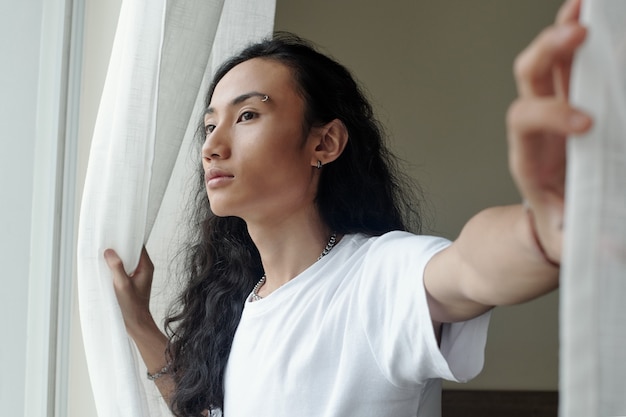 Hermoso joven en camiseta blanca abriendo cortinas en su dormitorio y mirando afuera