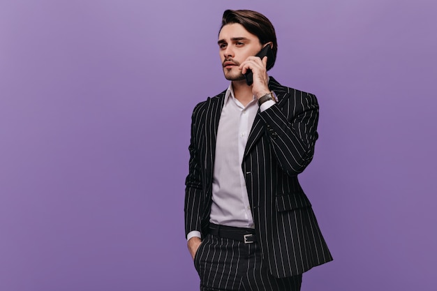 Hermoso joven empresario con cabello castaño, camisa ligera y traje a rayas de moda, hablando por teléfono y mirando a otro lado