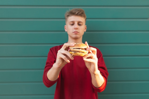 Hermoso joven se encuentra en la pared de color turquesa y tiene una hamburguesa apetitosa.