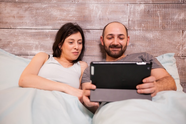 Hermoso joven esposo y esposa en pijama riendo mientras usa la tableta antes de acostarse.