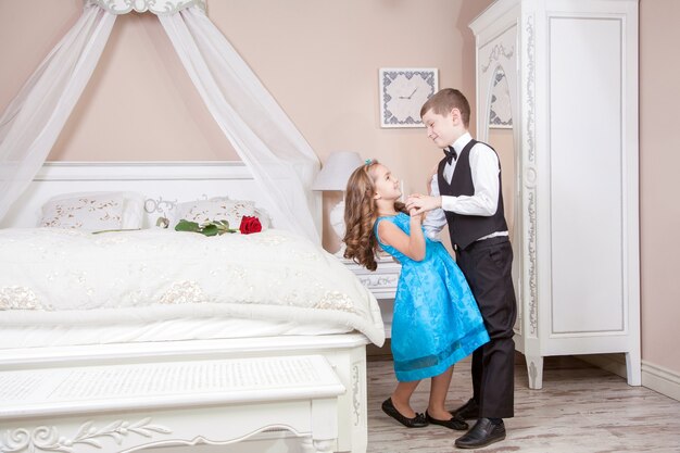 Hermoso joven príncipe en traje de esmoquin y princesa sosteniendo rosa en vestido azul y banda de moda posando sentada en la cama en una habitación luminosa y mirando a lo lejos. Historia de amor. Tiro del estudio.