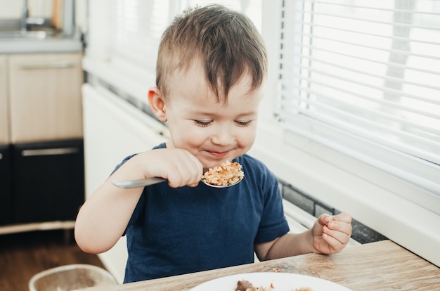 Hermoso lindo bebé come arroz con una cuchara en la cocina