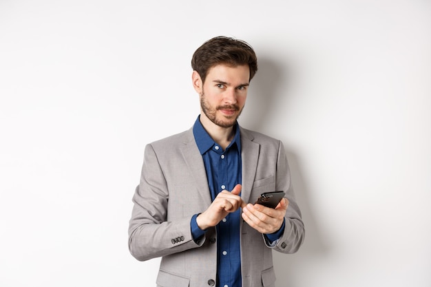 Hermoso modelo masculino barbudo en traje mediante teléfono móvil, sonriendo complacido a la cámara, fondo blanco.