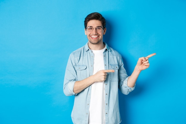 Hermoso modelo masculino caucásico con gafas, señalando con el dedo directamente a su logotipo, mostrando copia espacio, de pie sobre fondo azul.