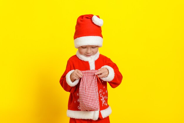 Hermoso muchacho caucásico de tres años vestido como Santa Claus abre un saco con papas fritas de año nuevo