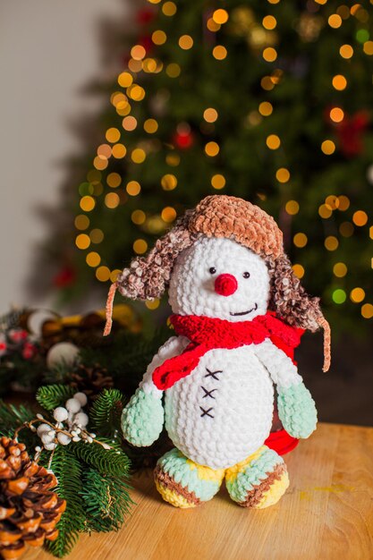 El hermoso muñeco de nieve de juguete tejido está parado sobre una mesa de madera El divertido juguete con cosas navideñas cerca