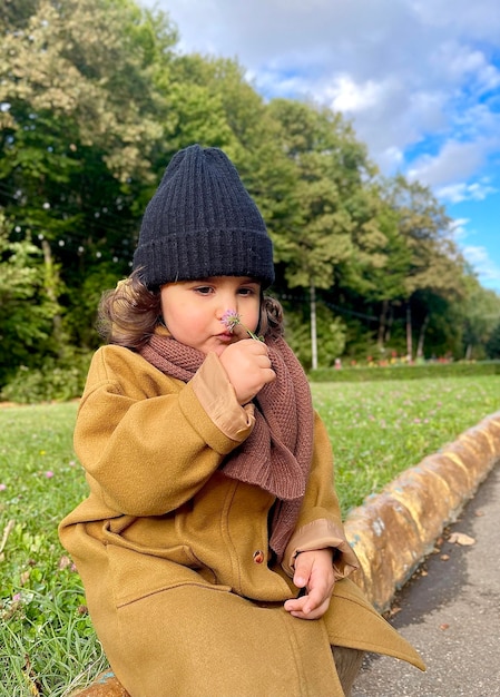 Un hermoso niño con abrigo y sombrero está sentado en la acera en un parque de otoño y olfateando una flor