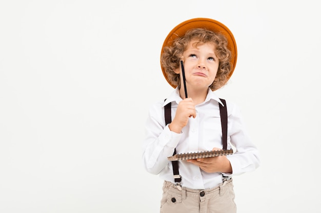 Hermoso niño con cabello rizado en camisa blanca, sombrero marrón, gafas con tirantes negros escribe en nota y piensa aislado en blanco