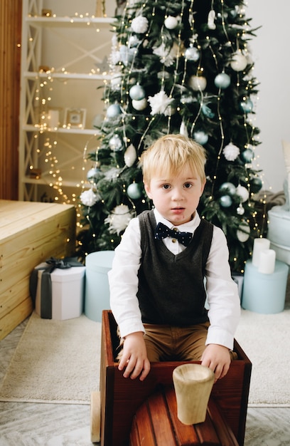 Hermoso niño caucásico sentado en un tren de juguete wodden delante de un lujoso árbol de Navidad