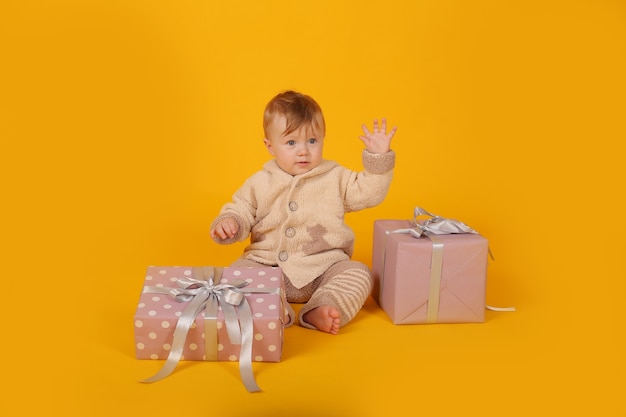 Un hermoso niño de ojos azules en un cálido traje de punto con cajas de regalo sobre un fondo amarillo